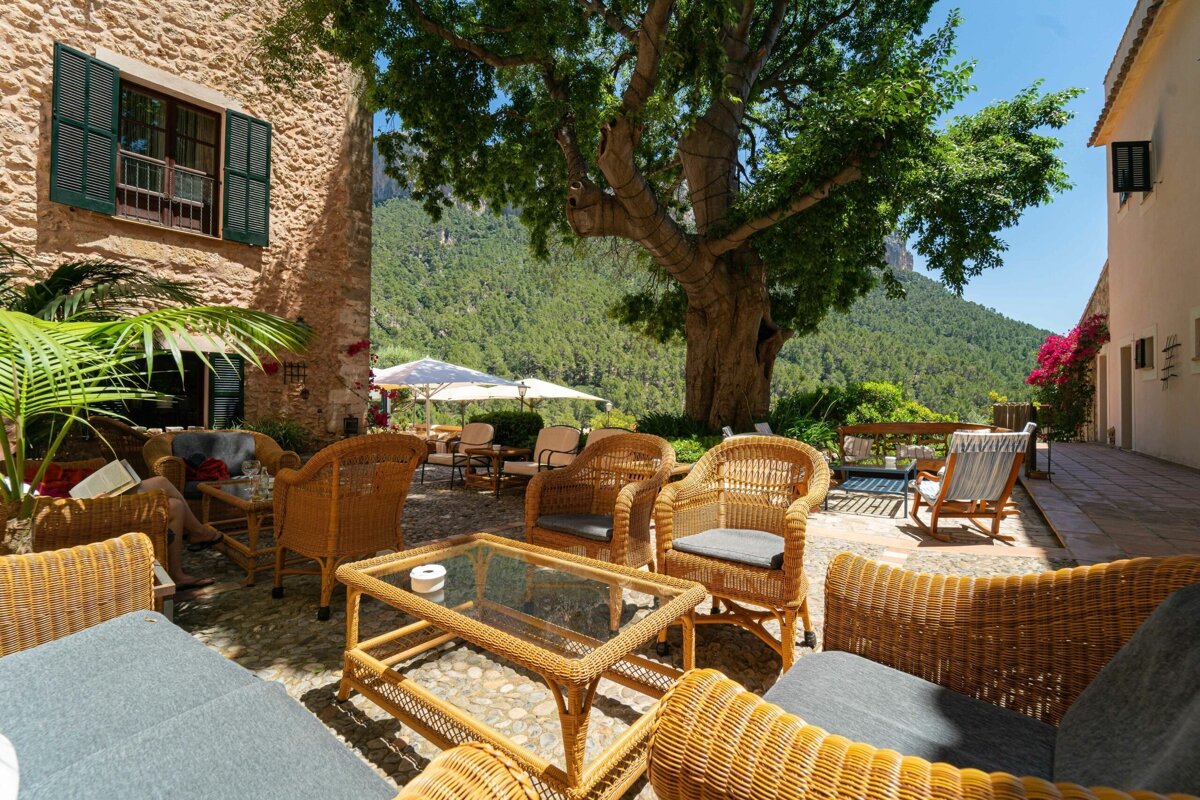 Sunny patio with wicker furniture, shaded by a large tree, next to a stone building with lush mountain views.