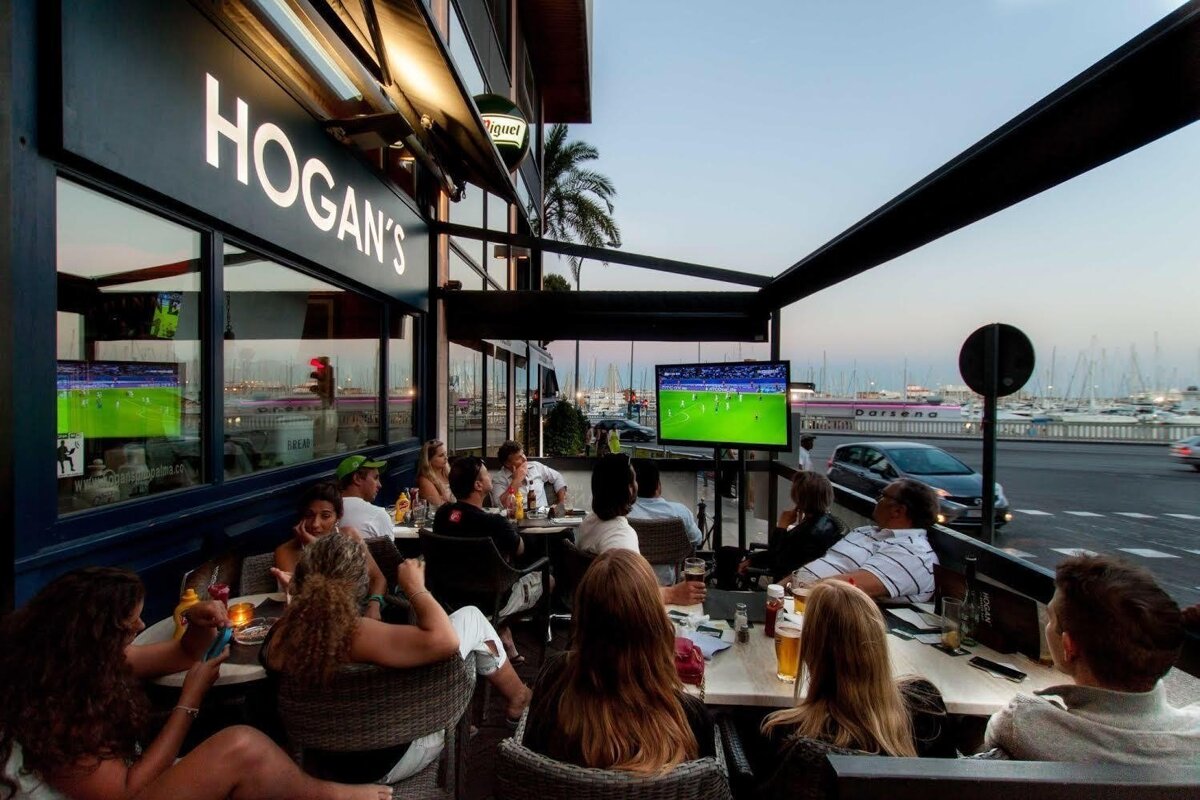 People watching a soccer game on TV outside Hogan's bar with a marina in the background at dusk.