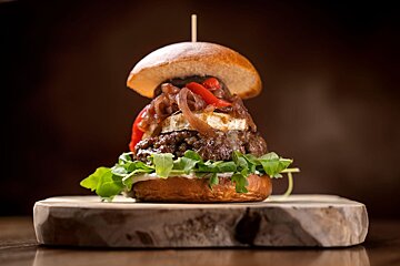 A gourmet burger with a thick patty, caramelized onions, red peppers, soft white cheese, arugula, and sauce, skewered on a wooden board.