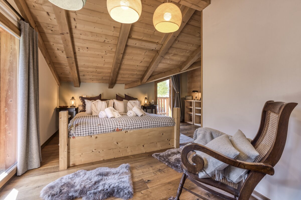 A cozy, rustic bedroom features a large wooden bed with plaid bedding, a beamed ceiling, and a vintage rocking chair. A door opens to a balcony.