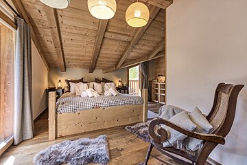 A cozy, rustic bedroom features a large wooden bed with plaid bedding, a beamed ceiling, and a vintage rocking chair. A door opens to a balcony.