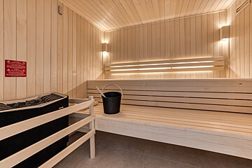 A bright, modern sauna with light wooden walls and benches. Features a black electric heater, bucket, ladle, and warm LED strip lighting.