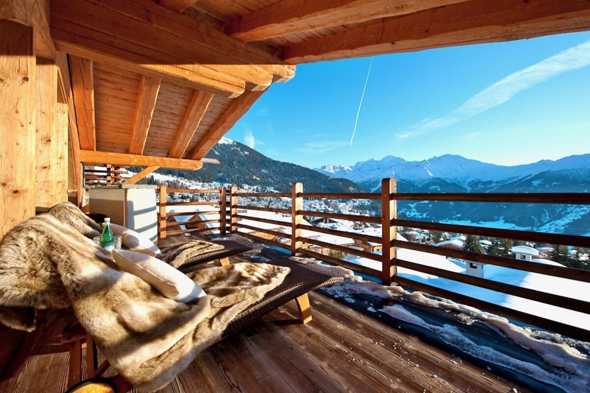 Cozy wooden balcony with fur blankets overlooking a snowy mountain village and majestic snow-capped peaks under a clear blue sky.