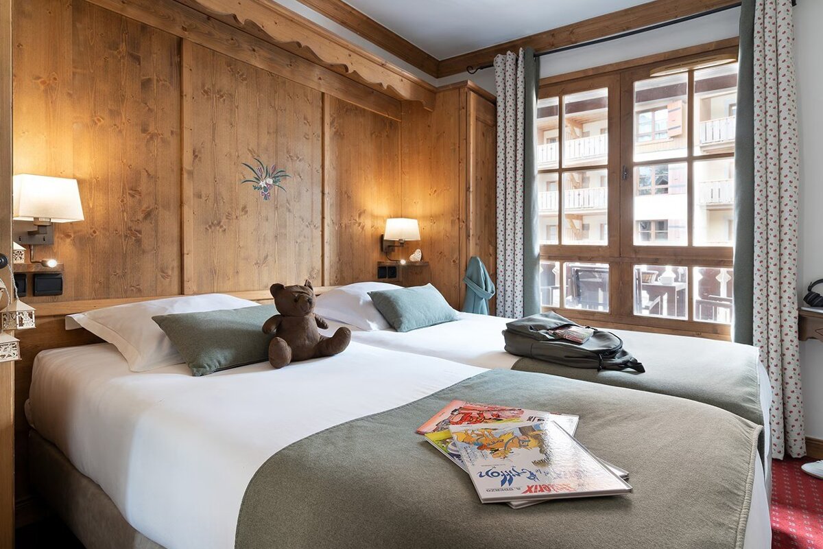 Cozy wood-paneled bedroom featuring twin beds with a teddy bear and magazines, next to a large window.