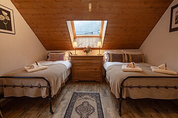 A cozy attic bedroom with two twin beds, a wooden ceiling, and a skylight. A dresser with lamps and flowers is positioned between the beds.