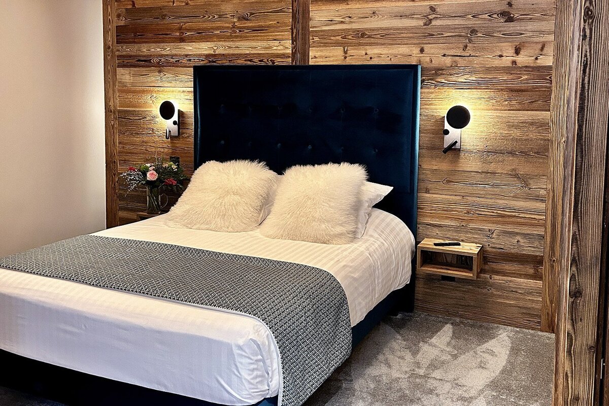 A stylish bedroom featuring a large bed with a dark blue headboard and fluffy pillows, set against rustic wood-paneled walls with modern sconces.