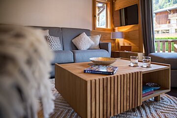 A warm, modern chalet living room with a gray sofa, wooden coffee table, and wood-paneled walls. A large window offers an outside view.