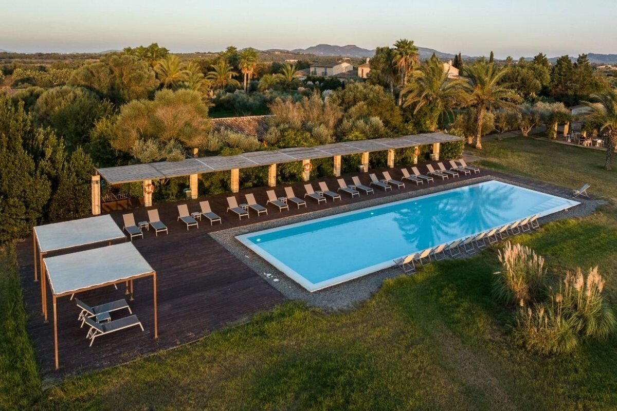 Aerial view of a long, clear blue pool lined with lounge chairs and pergolas, set amidst lush green trees and fields under a warm sky.