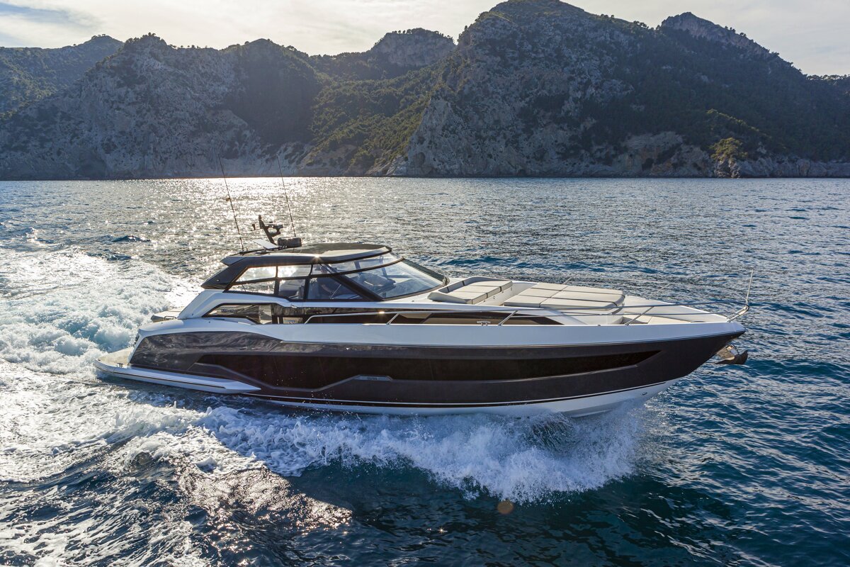 A modern yacht speeds across blue water, creating a white wake, with sunny mountains in the background.