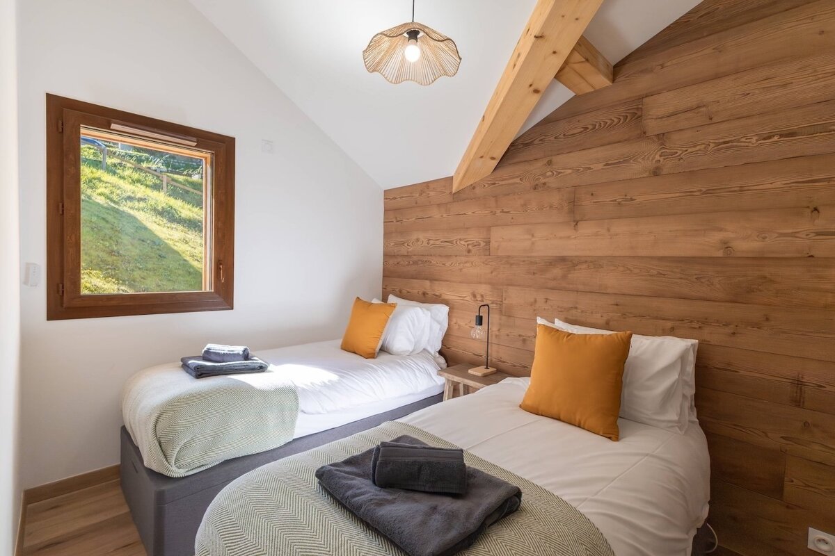 A rustic-modern twin bedroom features a wooden accent wall, white walls, and a bright window. Beds have white linens, orange pillows, and patterned throws.