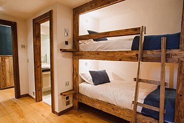 A cozy room features rustic wooden bunk beds with white bedding and blue accents, warm wooden floors, and white walls. Two doorways are visible.