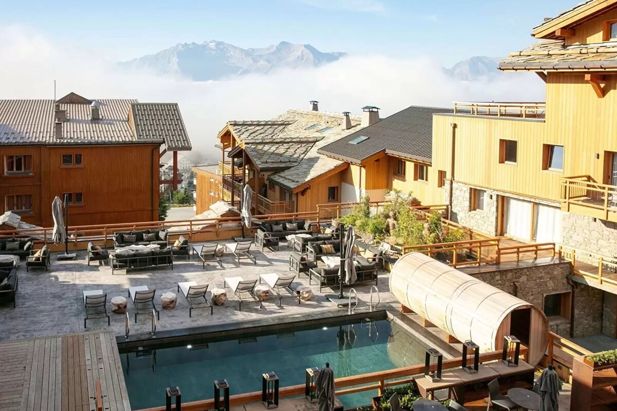 A scenic mountain resort with wooden chalets, an outdoor pool, lounge furniture, and a barrel sauna, overlooking cloudy peaks.