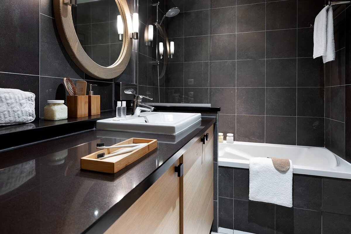 Modern bathroom with dark grey tile walls, a light wood vanity, round mirror, white sink, and a bathtub with fresh towels.