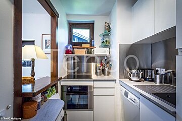 A compact, modern kitchen featuring white cabinets, gray countertops, an oven, dishwasher, microwave, and a window. A bar area with a lamp is visible.