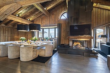 Luxurious rustic wooden chalet interior with a dining table set, a cozy fireplace with a TV, and panoramic mountain views from the windows.