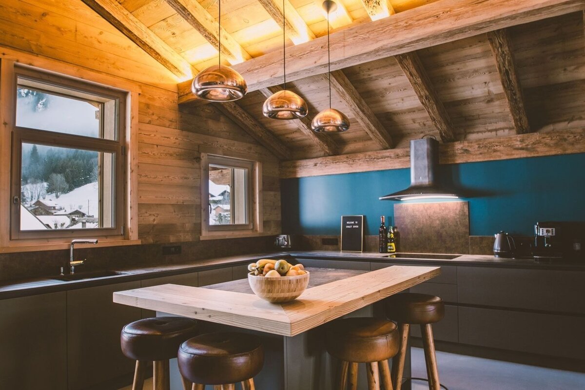 A stylish, modern chalet kitchen features rustic wooden beams, a teal accent wall, and an island with stools, offering views of snowy mountains.