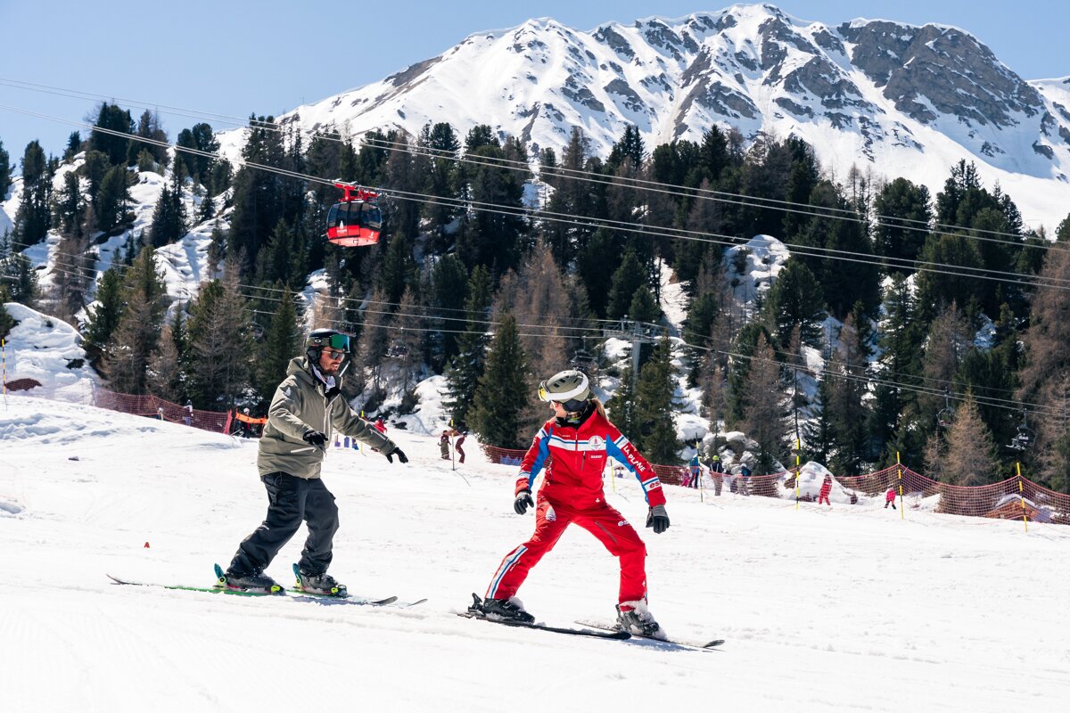 Two skiers, one in red, are on a sunny mountain slope. A red cable car moves overhead with pine trees and snowy peaks in the background.