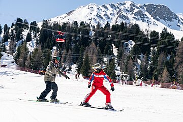 Two skiers, one in red, are on a sunny mountain slope. A red cable car moves overhead with pine trees and snowy peaks in the background.