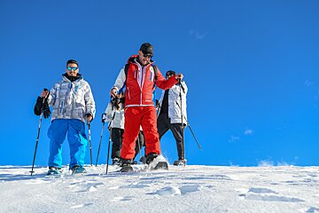 Several people in winter gear snowshoeing up a snowy slope against a vibrant blue sky, with some visible footprints in the snow.