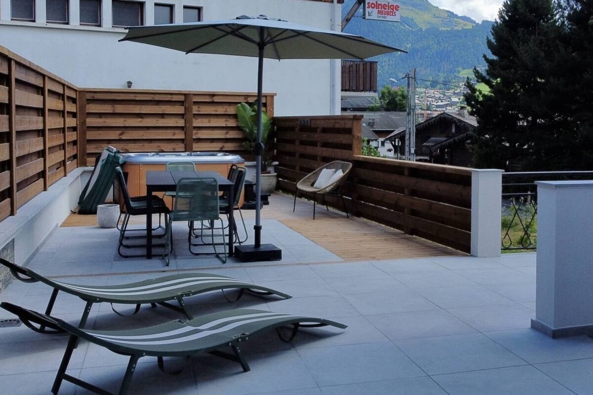 A modern terrace with a hot tub, outdoor dining set, loungers, and umbrella, featuring a mountain village view in the background.
