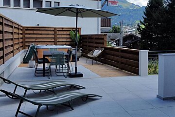 A modern terrace with a hot tub, outdoor dining set, loungers, and umbrella, featuring a mountain village view in the background.