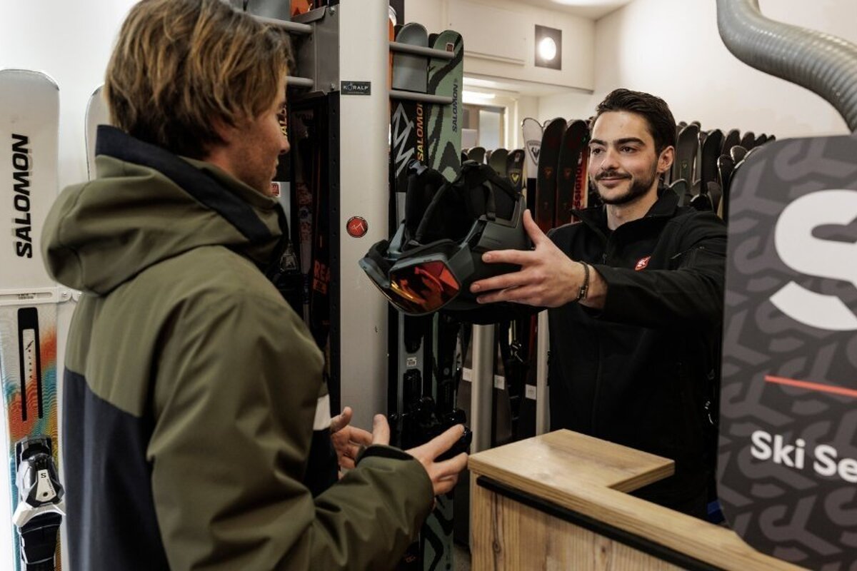A ski shop worker hands a helmet with goggles to a customer in front of racks of skis.