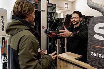 A ski shop employee hands a helmet with goggles to a customer in front of racks of skis.
