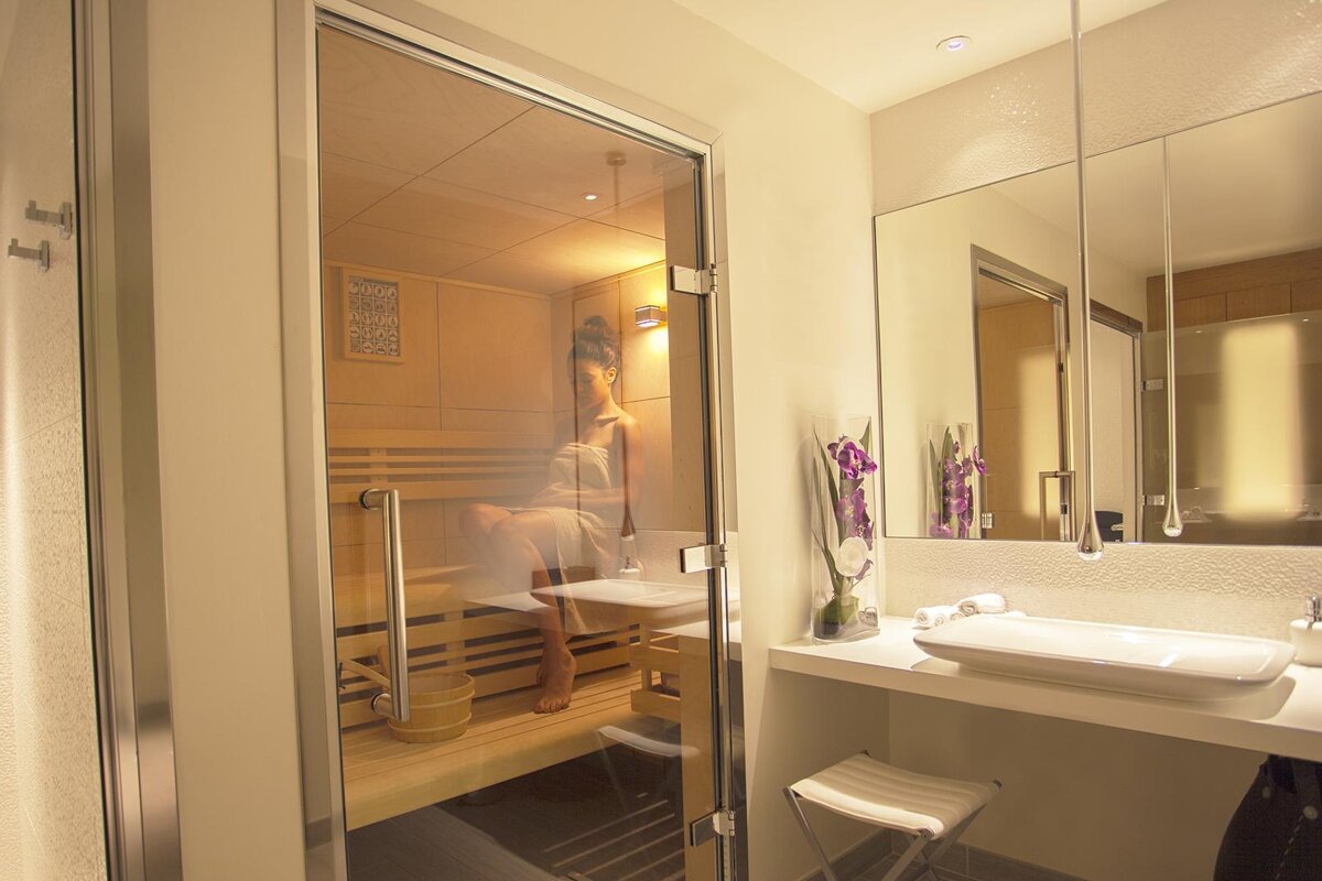 A woman sits in a sauna in a bathroom next to a sink