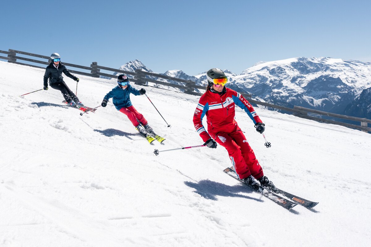 Three skiers, including a child, carve down a sunny, snowy mountain run with distant peaks under a clear blue sky.