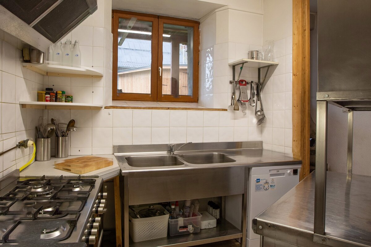 A functional kitchen with stainless steel counters, a gas stove, white tiled walls, a wooden window, and shelves with utensils and spices.