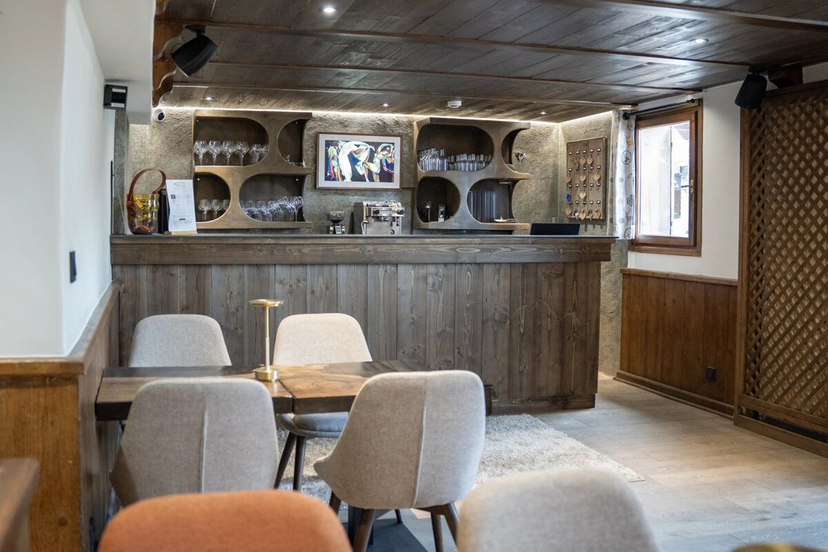 An inviting rustic lounge/bar features warm wood paneling, a stylish counter with glass displays, a coffee machine, and modern cushioned chairs around a table.