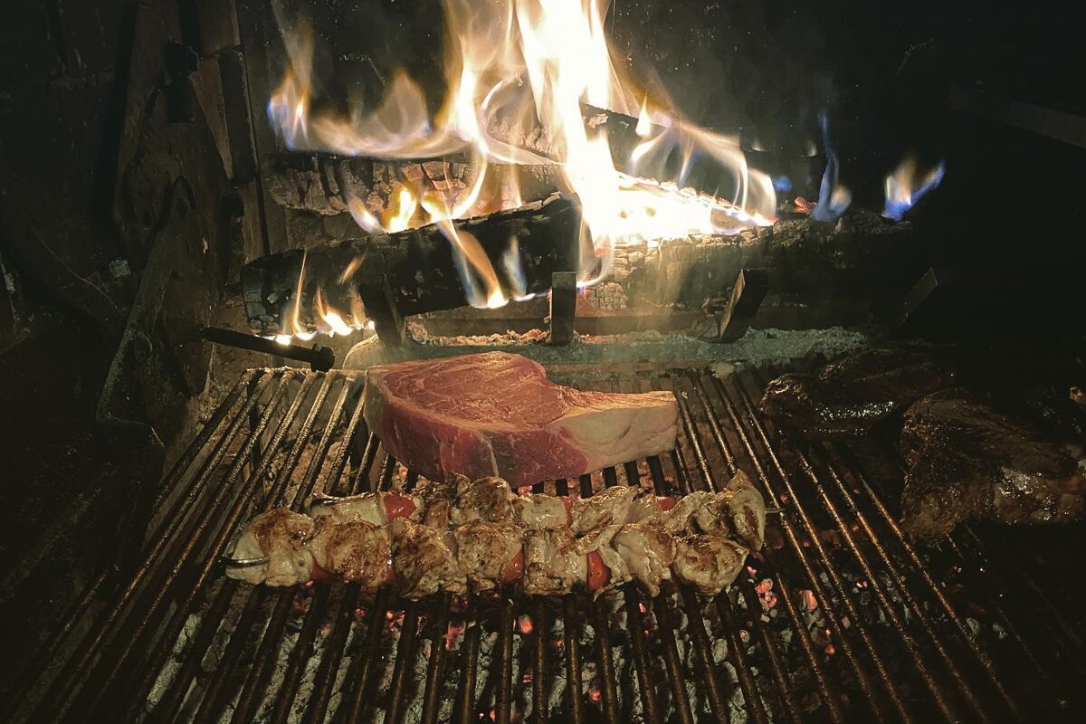 Steaks and shish kebabs are cooking on a grill in front of a fire