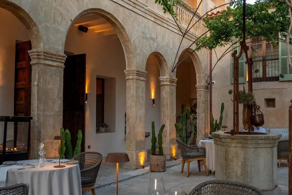 An inviting outdoor restaurant in a stone-arched courtyard, with set dining tables, warm wall and floor lighting, and scattered cacti.