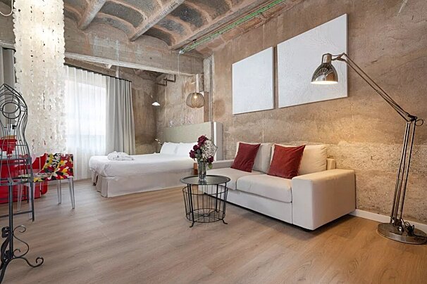 A stylish industrial studio featuring a white bed and sofa, concrete walls, light wood floors, and modern furnishings.