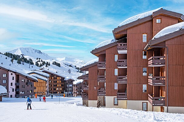Skiers are skiing down a snowy slope in front of a building with balconies
