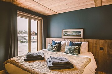 Cozy bedroom with a dark blue accent wall, wooden ceiling/headboard, and a large window offering a view of a snow-covered village.