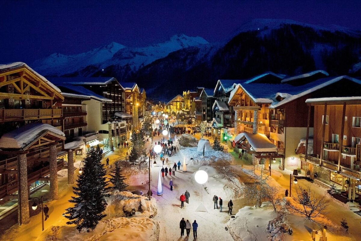 A beautiful snowy mountain village lit up at night, with chalet-style buildings, people walking, and snow-capped peaks in the background.