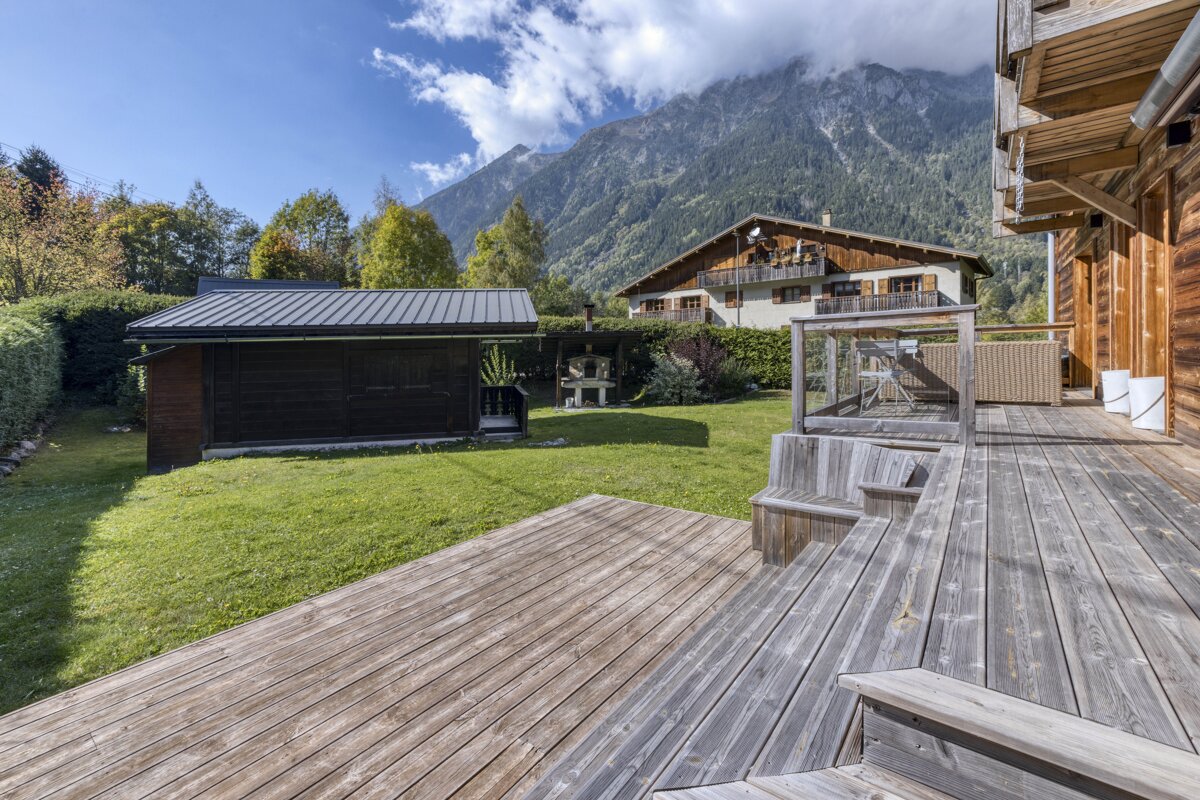 A sunny mountain chalet backyard with a wooden deck, green lawn, shed, and a distant house against a backdrop of cloudy peaks.