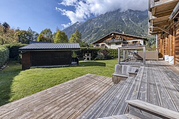 A sunny mountain chalet backyard with a wooden deck, green lawn, shed, and a distant house against a backdrop of cloudy peaks.