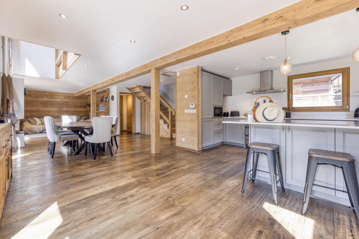 A spacious, open-plan living area features a dining table, a modern kitchen with a grey island, and a lounge area. Wooden floors and exposed beams create a cozy, rustic feel.