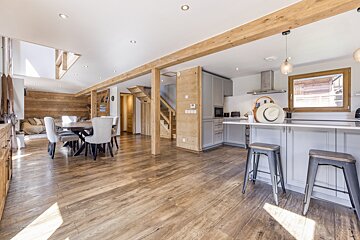 A spacious, open-plan living area features a dining table, a modern kitchen with a grey island, and a lounge area. Wooden floors and exposed beams create a cozy, rustic feel.