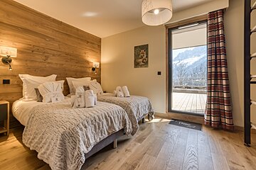 Chalet-style bedroom featuring two twin beds, wood paneling, bathrobes, and a balcony overlooking a snowy mountain range. Fox art on the wall.