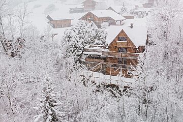 Snow-laden wooden chalets nestled in a frost-covered forest during a heavy winter snowfall, creating a serene, picturesque scene.
