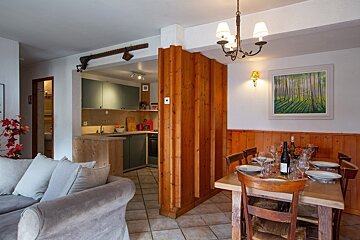 An open-plan living, dining, and kitchen area. Features a grey sofa, a rustic wooden dining table set for guests, and a modern kitchen with green cabinets. Warm wood accents throughout.