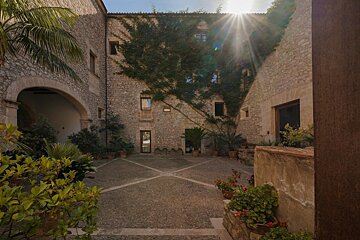 A sun-drenched, rustic stone courtyard with ivy-covered walls, potted plants, and an archway. Bright sun rays gleam over the old architecture.