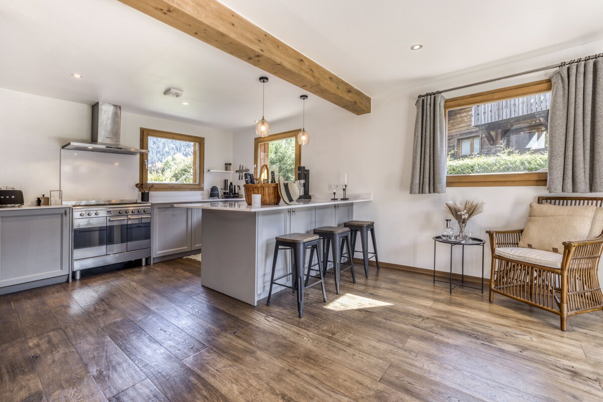 A bright, modern kitchen with light grey cabinets, a large island, warm wooden floors and ceiling beam. Windows provide views; a rattan chair adds coziness.