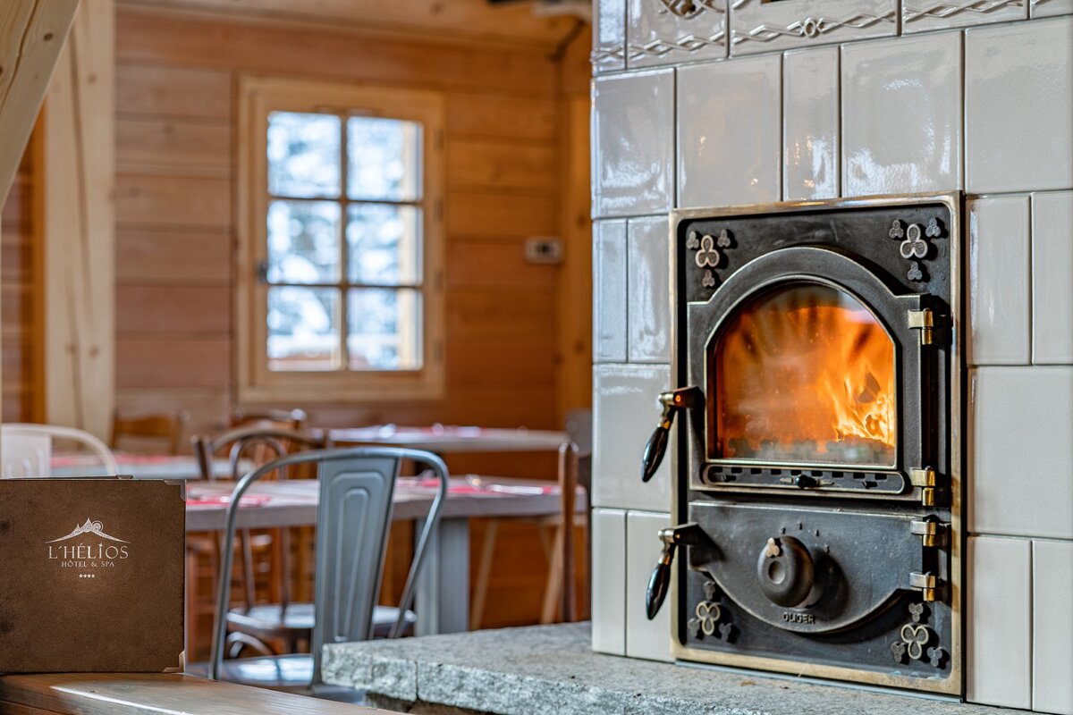 A cozy, rustic hotel interior features a brightly burning tiled fireplace, wooden walls, a snowy window view, and an L'Hélios menu.