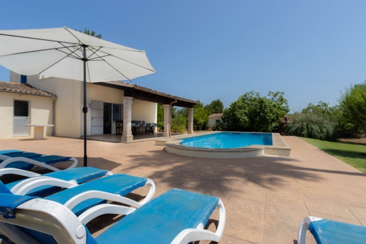 A sunny villa with a patio, blue lounge chairs, a white umbrella, and a sparkling swimming pool, surrounded by green landscaping.