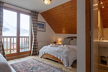 Cozy bedroom with a balcony overlooking foggy mountains, featuring wood paneling, a patterned rug, and a peek into an en-suite bathroom.