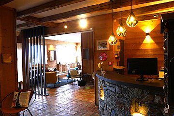 A warm, rustic hotel lobby with wood paneling, exposed beams, a stone reception desk, modern pendant lights, and a cozy seating area in the background.